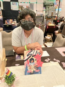 A photo of Aatmaja Pandya wearing a mask and holding a copy of Slip. The book has stickers saying Sold Out - More Tomorrow
