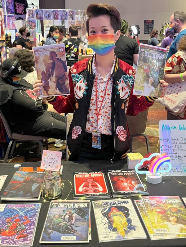a photo of Alyssa Wong wearing a rainbow mask and holding up Doctor Aphra comics