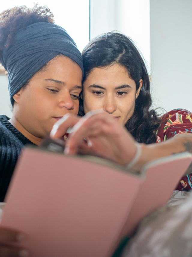 two women, one with light brown skin and one with pale skin, reading a book together